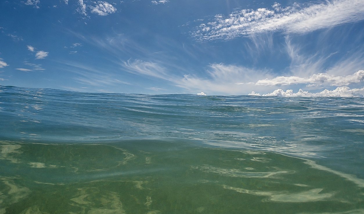 O mar de Iracema visto de dentro da água, rente à superfície, com o horizonte no alto e nuvens ao fundo.